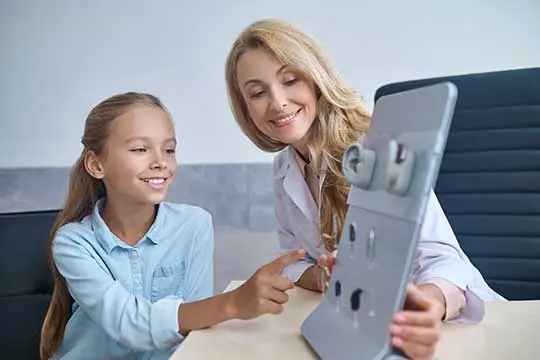 A young girl and a smiling woman look at and discuss various hearing aids displayed on a stand in a medical office. The girl is pointing at the hearing aids while the woman holds the display.