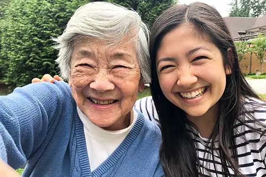 Two women, one older and one younger, are smiling and taking a selfie together outdoors. The older woman has gray hair and wears a blue sweater, while the younger woman has long dark hair and wears a striped shirt.