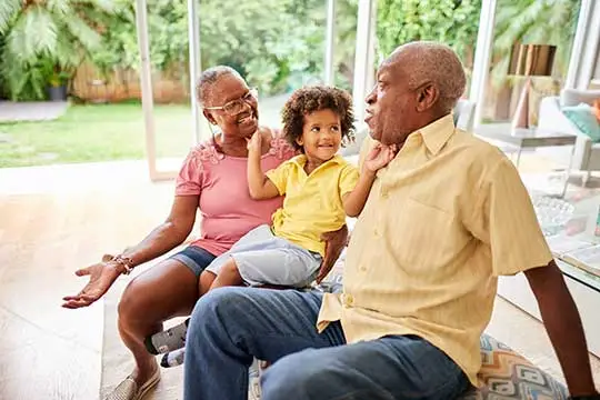 An elderly couple sits inside by large windows, smiling and talking with a young child who sits on their laps. The background shows a green garden outside. All three appear happy and engaged.